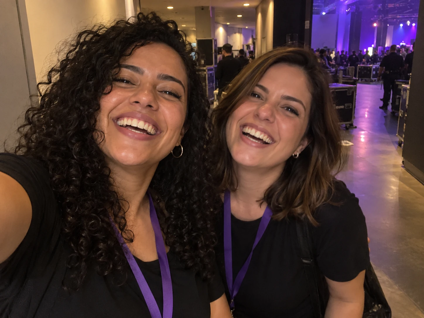Selfie ilustrativa de duas amigas sorrindo no corredor de um evento.